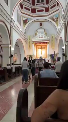 Inside a church, a line of people moves down the aisle toward the altar.  The church has a white ceiling and white walls with archways and paintings.  Banners that read “SAN CALIXTO” and “PROTEGENOS” hang on either side of the altar, which features a large gold crucifix.  Pews line both sides of the aisle, and some people are sitting in them.  Most of the people in the line are adults, though one young child wearing a pink shirt and white pants walks hand-in-hand with an adult woman.  The floor is patterned with red and white designs.  The camera is positioned at a slight angle from one of the pews.