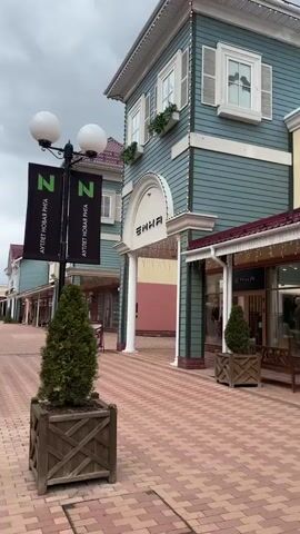 The camera pans across an outdoor shopping center. The ground is paved with red and pink bricks. On either side are two-story buildings with red roofs and pale blue siding. Some of the buildings have white columns and arched doorways. There are store signs over the doorways. In the foreground is a light post with two spherical lights. Hanging from the post are two black banners with the words "АУТЛЕТ НОВАЯ РИГА" written in white. A small, evergreen tree in a wooden planter stands next to the post. The sky is overcast. There are no people visible in the video.