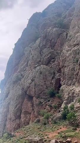 A small, dark drone ascends from behind a large, reddish-brown rocky cliff face on the left side of the frame. The drone continues to ascend, moving into the center of the frame against a cloudy sky with grey and white clouds, and then hovers. The rocky cliff, partially covered with sparse green vegetation, remains visible on the right side of the frame throughout the drone's flight.