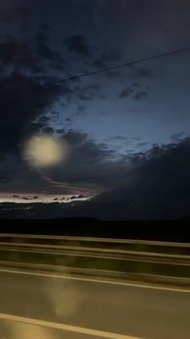 A video captures a view from a moving vehicle at dusk. The sky is filled with dark, dramatic clouds, with hints of pink and orange light peeking through the horizon. Below the sky, a dark, silhouetted landscape of hills or mountains is visible. In the foreground, there are visible traffic barriers or guardrails, indicating the vehicle is on a road, possibly a highway, with lane markings suggesting multiple lanes of travel. The overall mood is atmospheric and potentially somber due to the dim lighting and dark clouds. There are no people or distinct objects visible within the frame besides the natural landscape and road infrastructure.