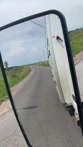 The video shows a first-person perspective from inside a vehicle, likely a truck, looking at the side-view mirror. The mirror reflects a winding asphalt road through a rural landscape with green grass and trees on either side. Power lines and poles are visible along the road. In the distance, there are some buildings and a body of water. The sky is partly cloudy. The vehicle appears to be moving forward.