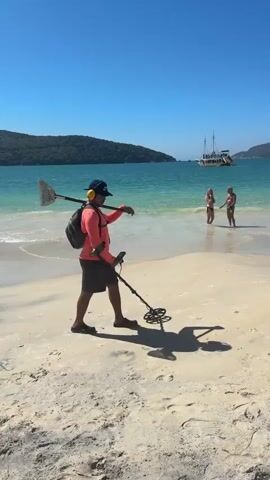 A man walks along a sandy beach on a sunny day, using a metal detector. He is wearing a coral-colored long-sleeved shirt, dark shorts, a blue baseball cap, and yellow ear protection. He carries a fishing net with a long handle over his right shoulder and has a backpack on. The metal detector is held in his right hand, with its coil near the sand. His shadow is visible on the beach. In the background, the ocean is turquoise and clear, with waves gently lapping the shore. Two women are standing in the shallow water further down the beach. A sailboat with sails is visible in the distance on the water, along with other boats. The coastline is lined with green hills and rocky outcrops.