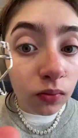 A close-up video shows a woman curling her eyelashes with a silver eyelash curler. She has light skin, dark eyebrows, and a nose ring. She is wearing a grey sweater and a sparkly necklace. Her eyes are partially open and focused on the curler. There is some ambient sound, perhaps from an airplane or bus.