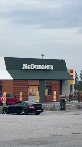 A black car is parked in front of a McDonald's restaurant. The McDonald's sign is visible at the top of the building. The restaurant has a drive-thru window. There are several parking spots marked with numbers. The weather appears to be overcast.
