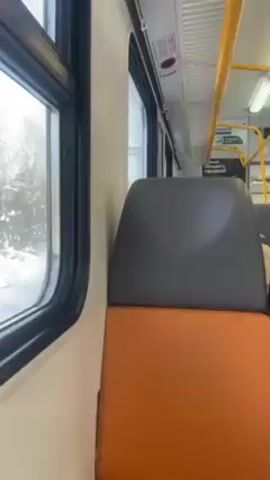 The video shows the interior of a train car. A window on the left side shows a snowy landscape passing by. There is an empty orange and grey seat in the foreground. Above the seat is a white wall with an overhead light. A yellow handrail is visible near the top of the frame. The train car appears to be in motion.