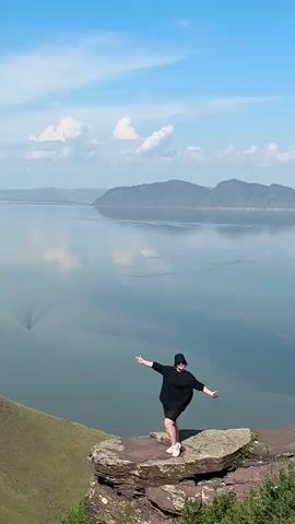 A person in a black shirt and shorts stands on a rock outcropping, arms spread wide. Another person, wearing a red cap and backpack, kneels on the grass below, holding a phone to their face as if taking a picture. The background features a large body of water, mountains, and a blue sky with white clouds. The scene appears to be a scenic overlook on a sunny day.