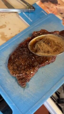 The video shows a close-up view of a seasoned steak on a blue cutting board. A spoon is being used to pour a liquid, likely pan juices or a sauce, over the steak. The steak appears to be well-cooked with a seared crust. The background is slightly blurred, but hints of a kitchen environment are visible, including what looks like a cookbook or magazine and kitchen utensils. The lighting is bright, highlighting the texture and sheen of the steak. The action is focused on basting the steak with the liquid.