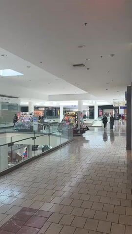 The video shows a view of a shopping mall interior, likely during the daytime given the bright, artificial lighting and natural light filtering in from skylights. The camera pans across a wide, tiled corridor. In the foreground, a glass railing overlooks a lower level of the mall, where more shops and escalators are visible.

Several people are walking through the mall. On the upper level, a few individuals are browsing at shops. A prominent display, featuring a variety of stuffed animals and toys, is situated near a pillar with the word "TIKTOK" printed vertically on it. Further down the corridor, two women are walking side-by-side. In the distance, more shoppers and store entrances can be seen.

The mall appears to be moderately busy, with a relaxed atmosphere. The overall setting is that of a typical indoor shopping mall. No specific time of day or weather conditions are evident beyond the general brightness. The visible text includes "TIKTOK," "AMERICAN EAGLE," and what appears to be the logo for "MAC Cosmetics." The location context provided is National City, United States of America.