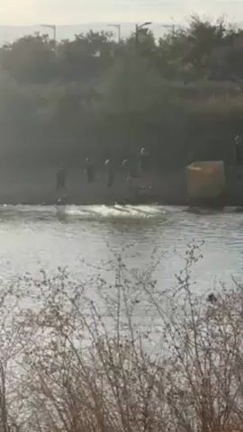 The video shows a group of people standing on the bank of a body of water. The group consists of 6-8 people, and they appear to be adults. They are positioned at the edge of the water, and some are near a yellow crate. The body of water is calm, and the bank is lined with vegetation. The background features more trees and possibly bushes, with a hint of sky in the distance. The video has a slightly hazy or muted quality.