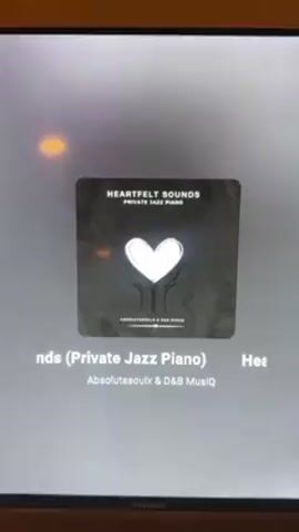 A close-up shows a TV screen displaying the album art for "Heartfelt Sounds: Private Jazz Piano" by Absolutesoulx & D&B Musiq. The album cover is dark gray with a textured, vintage look. At the center is a white heart held up by two hands. A Sharp logo is visible at the bottom of the TV. The music is playing.