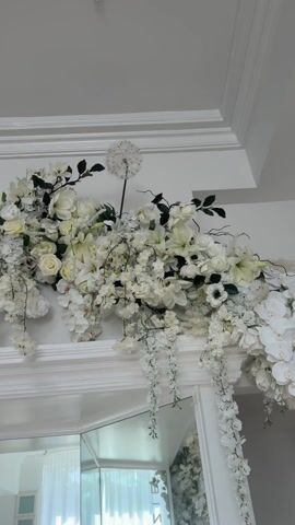 This video presents a static, eye-level shot of an opulent white floral arrangement situated above a mirrored surface in an upscale interior. The elaborate display features a rich assortment of artificial and possibly fresh flowers, predominantly in shades of white, including roses, orchids, lilies, cherry blossoms, dahlias, and magnolias. Subtle accents of pale pink or peach blooms and lush green foliage are interspersed throughout, creating a grand and cascading effect. A distinctive white, spherical, dandelion-like flower stands out at the top of the arrangement.

Below the floral display, a white, ornate mantelpiece-like structure with decorative crown molding is visible. The lower part of the frame reveals a large mirrored surface, reflecting elements of the room, such as a window or doorway draped with sheer white curtains, suggesting a bright and airy space. A small, black, circular device, possibly a camera or sensor, is discreetly mounted on the wall behind the flowers.

The overall aesthetic conveys a sense of luxury, elegance, and celebration, typical of event decor or high-end interior design. The bright, even lighting indicates an indoor setting, likely during the day or in a well-illuminated evening space. This scene is set in Beverly Hills, United States of America, aligning with the sophisticated and grand nature of the decor. No people are visible, and there is no discernible activity or text within the frame.