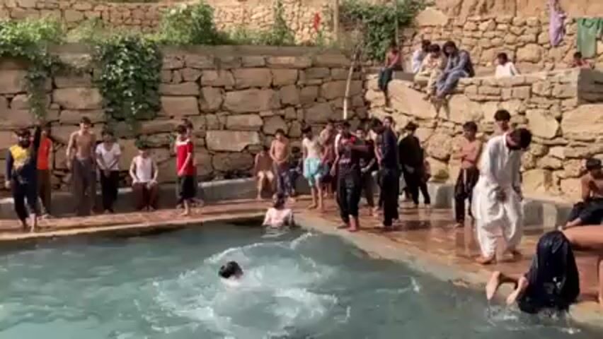 A group of about 30 people, mostly male children, are gathered around a rectangular outdoor pool. Some are in the water, swimming or splashing, while others are standing on the pool's edge. The pool has stone walls on either side, and some people are sitting on top of one of the walls. The people are dressed in a variety of casual clothing. Some are shirtless, while others are wearing shirts, pants, or traditional Pakistani shalwar kameez. The environment is outdoors with trees and vegetation in the background, suggesting a natural setting. The pool's water is clear and light blue.