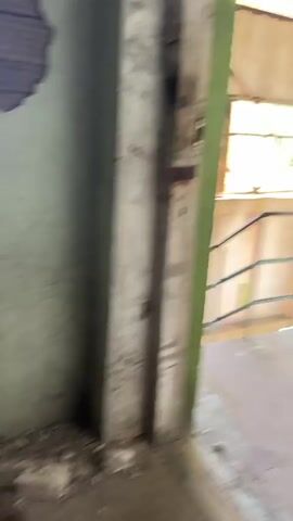 This video captures the interior of a large, abandoned industrial building, likely a factory or warehouse, located in Tsitrusovyj, Abkhazia. The scene is one of decay and disuse. The subjects are primarily the building itself and its contents.

The building has a high, sloped roof constructed of corrugated metal, supported by a metal frame. Natural light streams in through large, grimy windows along the sides and at the far end, suggesting it's daytime with bright, possibly sunny, weather. The walls are rough and stained, with some evidence of peeling paint. A metal railing and a concrete walkway are visible in the foreground, indicating a raised viewing platform or mezzanine.

The main open space of the building is filled with debris, dust, and concrete on the floor. In the center of the space, towards the back, is a large piece of industrial equipment, possibly a generator or a large machine, covered in red graffiti. Other smaller, unidentifiable objects and remnants of industrial activity are scattered around. Vegetation is creeping in from the outside, with some plants growing on the walls and near the windows.

There is no visible activity or event taking place; the atmosphere is still and silent, evoking a sense of abandonment and neglect. The overall emotion conveyed is one of disuse and the passage of time. No discernible text is visible in the video. The location cues, such as the architecture and the surrounding vegetation visible through the windows, along with the provided context, point to an abandoned industrial site in Tsitrusovyj, Abkhazia.