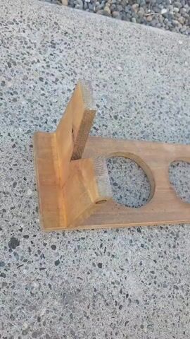 A wooden object with two circular cutouts and two upright wooden pieces is displayed on a concrete surface. The object appears to be a holder or stand, possibly for cups or bottles, as evidenced by the circular openings. The wooden material is a light brown color and shows some signs of wear and tear, with a rough texture in some areas. The object is positioned on a grey concrete surface that is dotted with small pebbles, suggesting an outdoor setting. The lighting suggests it is daytime. The video captures the object from various angles as it is rotated and moved. The camera's focus shifts between the wooden object and the surrounding environment, with the pebbles and concrete being clearly visible. The overall presentation is simple and direct, showcasing the object without any additional context or embellishments.