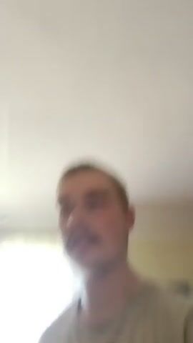 The video features a close-up, somewhat blurry shot of a young man. He is wearing a light-colored t-shirt. The man is looking directly at the camera with an open mouth and his tongue sticking out, creating a playful or surprised expression. In the background, there is a window with sheer curtains, allowing bright light to filter through, suggesting it's daytime. Behind him, a television screen is visible, displaying what appears to be a colorful image or video. The room seems to be indoors, possibly a living room or bedroom, with a ceiling light fixture also visible in the upper left corner of the frame. There is no visible text in the media file. The overall mood is informal and candid.