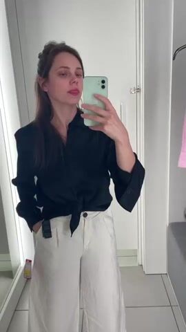A woman stands in a fitting room, taking a selfie with her phone. She is wearing a black, tied-up shirt and white, wide-leg pants. She has her hair pulled up in a bun. The fitting room has bright lights, a mirror, and a bench.