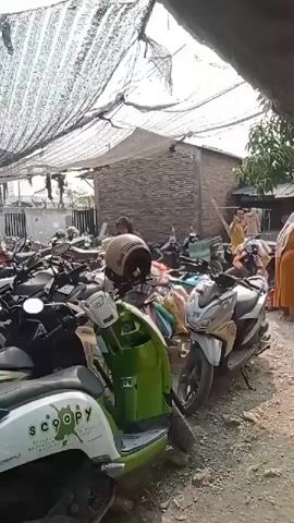 The video shows an outdoor scene with many motorcycles parked under a netting structure. Several people are visible, some are interacting with each other, possibly in a marketplace setting. One person in an orange dress is handing something to another person. A green and white scooter is in the foreground. The ground is dirt and gravel. The overall impression is a busy, somewhat cluttered outdoor area.
