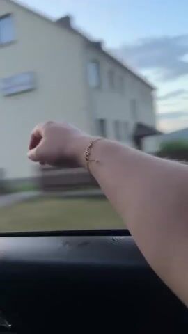 A close-up shot shows a person's right arm and hand with a gold bracelet on the wrist extending out of a car window. The arm is positioned in the foreground against a blurred background of a residential area with houses and trees. The person's hand is moving, and the fingers are slightly splayed. The camera is looking out from inside the car, with the car's interior visible at the bottom of the frame. The lighting suggests it is either early morning or late afternoon, with soft, natural light.