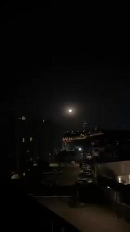 The video shows a nighttime scene of a city under a bright full moon. The foreground features a parking lot filled with cars and the silhouette of a building. In the background, a city skyline is faintly visible with streetlights lining the roads. A tall, lighted structure rises above the city buildings. The sky is dark, with the prominent moon as the main source of illumination.