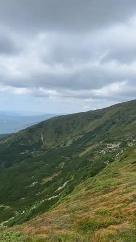 The video shows a panoramic view of mountains. The mountains are covered in green vegetation, with some patches of grass and trees. The sky is cloudy, with patches of blue sky visible. The camera pans across the landscape, showing different angles of the mountains and the valleys between them. The scene is serene and peaceful, with a sense of vastness and natural beauty.