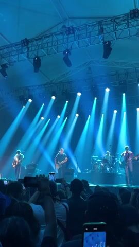 The video shows a concert from the perspective of someone in the audience. A band of four musicians is on stage. They all appear to be playing instruments, including guitars and drums. Bright blue spotlights shine down on the stage and the band. In front of the stage, a crowd of people is visible, with many holding up their phones to record the performance. The stage has a truss structure overhead, supporting the lighting. The environment appears to be an indoor venue with a tent-like structure above.