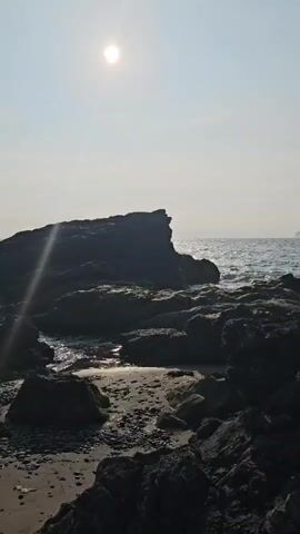 The video shows a beach scene with rocks and the ocean. It appears to be a picnic or camping setup. In the foreground, there is a portable grill with two skewers of meat cooking over coals. A bottle of sauce and a grill rack are also visible nearby. The sun is shining, casting a glare in the sky. The overall impression is a relaxed outdoor meal by the sea.