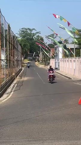 A person rides a red and white motorcycle down a paved road. They are wearing a black hijab and a pink and purple outfit. They are on the right side of the road. There is a tall chain-link fence on the left side of the road with a rusty metal structure behind it. On the right side of the road, there is a pink wall and several colorful flags are hanging from poles along the road. Trees and buildings can be seen in the background. The sky is blue and the weather appears to be sunny.