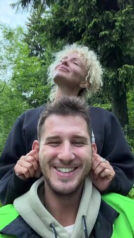 A medium shot shows a man with short brown hair and a trimmed beard smiling at the camera. He wears a light grey hooded jacket with bright green shoulder detail. Behind him stands a woman with curly blonde hair wearing a black sweatshirt. Her eyes are closed and she seems to be enjoying the sunshine. She is pulling on the man’s ears. They are in a forest setting with trees in the background.