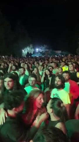 The video shows a crowded outdoor music concert at night. The audience is densely packed, with many people visible in the foreground and extending into the background. Lights, likely stage lighting, illuminate the crowd with green and yellow hues, casting shadows and creating a dynamic atmosphere. Many individuals are holding up their cell phones, presumably to record or photograph the event. Some people are raising their hands in the air, indicating enjoyment and participation in the music. Trees and greenery are visible in the background, suggesting the concert is taking place in a park or natural setting. There are also glimpses of tents or canopies in the distance. The overall scene depicts a lively and energetic gathering of people enjoying a live musical performance.