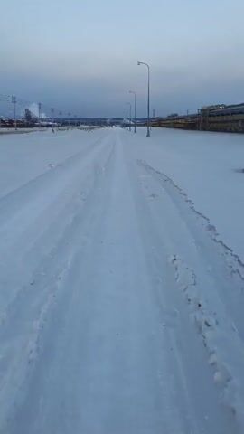 The video shows a snow-covered road leading away from the camera. Tire tracks are visible in the snow. On either side of the road are industrial structures. Several lampposts line the side of the road. The sky is overcast. There is a faint hum audible in the background. The camera appears to be moving forward. Footprints appear in the snow on the right-hand side of the road.