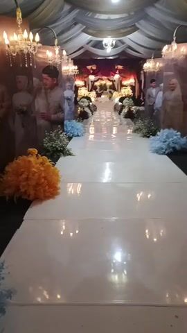 The video shows a decorated aisle, likely for a wedding or formal event. The aisle is covered in white fabric and lined with white flowers and blue and orange floral arrangements. Large portraits of a couple are visible on either side of the aisle. The ceiling is draped with fabric, and chandeliers hang from above. The overall impression is one of elegance and celebration.