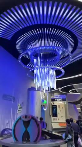 The video shows a modern, futuristic-looking interior, possibly an exhibition or museum. The dominant feature is a large, elaborate light fixture hanging from the ceiling. The fixture consists of concentric circles of lights with vertical LED strips hanging down, all glowing in bright blue. The ceiling above has a circular pattern of white lights radiating outwards.

Below the light fixture are several interactive exhibits. One appears to be a large, rounded machine with a screen displaying a sand timer design. Another exhibit has a screen with images and text, possibly providing information about the displayed content. Several people are present, including children, who appear to be interacting with the exhibits. They are dressed casually. The overall environment is well-lit with a cool, blue hue.
