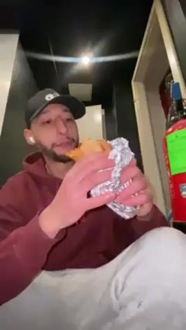A man wearing a maroon hoodie, light gray sweatpants, and a dark baseball cap is sitting on the floor in a narrow hallway. He has a beard, and he is wearing wireless earbuds. He is unwrapping and eating something wrapped in aluminum foil, presumably a sandwich or burrito.  To his right is a doorway to a storage area, in which there is a fire extinguisher and a bottle of wine. The hallway has dark walls and a light-colored ceiling.  The camera angle is from below.