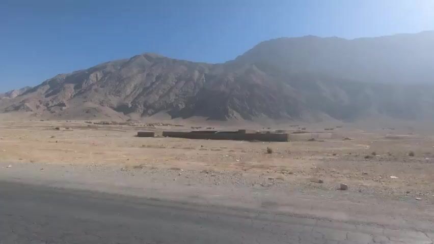 The video depicts a desolate, arid landscape, possibly a desert or mountainous region. The foreground features a paved road with visible cracks and fissures. Beyond the road, the terrain is dry and barren, with sparse vegetation. In the mid-ground, there are a few small, brick structures or ruins scattered across the landscape. A large, rocky mountain rises in the background, partially obscured by haze or dust. The sky is clear and blue. The overall impression is one of remoteness and aridity. The camera appears to be in a moving vehicle.