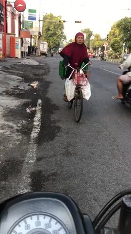 A woman wearing a maroon headscarf and dress rides a bicycle on an asphalt road. She is carrying a green basket on the handlebars and a white bag behind the seat. The road appears to be wet in places, with some debris scattered on the left side. In the background, there are buildings, traffic lights, trees, and other vehicles, including a motorcycle to the right. The sun appears to be low in the sky, suggesting it may be early morning or late afternoon. The video is shot from the perspective of someone on another moving vehicle, likely a motorcycle, as evidenced by the dashboard visible at the bottom of the frame.