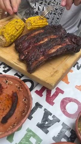 The video shows a close-up of a wooden cutting board on a table with a gray-shirted person holding a fork near two grilled corn cobs and a slab of barbecued ribs. A small bowl with food is partially visible in the lower left corner. The table is covered with a patterned tablecloth. The person appears to be holding one of the corn cobs with the fork.