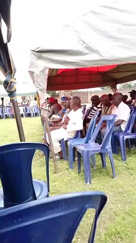The video shows an outdoor gathering of people seated on blue plastic chairs under a tent. The tent has a white tarp and a red lining. The ground is covered with grass. In the foreground, two blue plastic chairs are visible from the back. In the middle ground, a group of men are seated, facing towards the left. One man in the center is wearing a white outfit and appears to be holding a stick. The man to his right is wearing a striped shirt. Further to the right, other men are seated. In the background, more people are visible and some structures can be seen behind the tent. The atmosphere appears calm and orderly.