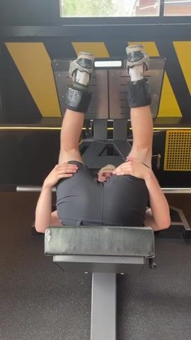 The media file shows a person lying on their back on a leg press machine in a gym. Their legs are extended upwards and their feet are placed on the footplate of the machine. They are wearing black shorts and white socks with black ankle weights. The person has blonde hair and is looking towards the camera. The gym has yellow and black striped walls and a grey floor. The time of day is not specified, but the lighting suggests it is daytime. The person appears to be performing a leg exercise.