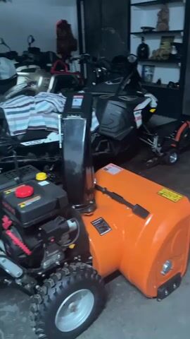 The video shows a cluttered garage. In the foreground, there is an orange snow blower, followed by a lawn mower. In the background, there are two snowmobiles, an ATV, shelves with various items, and a door leading to an outdoor area.
