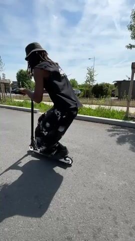 A person is riding a kick scooter on an asphalt road on a sunny day. The person is wearing a black sleeveless shirt, patterned shorts, black socks, and black shoes. They also appear to be wearing a helmet. The scooter is black with small wheels. The person is riding away from the camera, and the background includes a grassy area with small trees, a fence, parked cars, and some buildings. The sky is blue with scattered clouds. The person has dark skin and long hair.