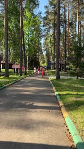A long asphalt path stretches away from the viewer, flanked by tall trees, forming a corridor-like view. A line of people, mostly wearing red and white clothing, is heading toward the end of the path. A young boy in a bright red hooded outfit runs toward the viewer on the path's right side, while another child in a yellow shirt and dark pants is on the path's center, pausing briefly to look directly at the camera. On either side of the path are green lawns and occasional wooden structures resembling shelters or small cabins. The path has a painted edge of green, yellow, and grey. The overall environment suggests a park or recreational area. The sky is blue and the lighting is bright, indicating daytime.