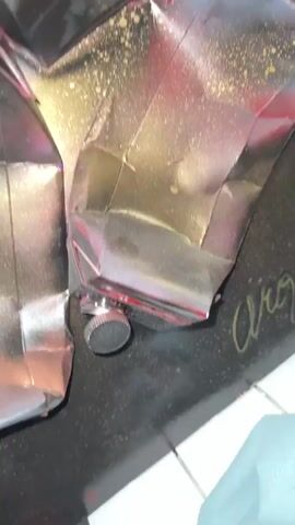 The video displays a close-up, shaky view of crumpled, metallic objects, possibly cans or wrappers, coated in silver, red, and gold spray paint. Some of the objects have visible lids or caps. The background is dark with some white and black elements suggesting a floor or surface. A faint yellow signature or marking is visible on the dark surface. The lighting is dim, and the focus shifts and blurs throughout the clip.