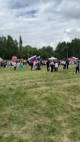 A group of people are gathered outdoors in a grassy field under a cloudy sky. Several flags, including the Russian flag, are visible. There are tents and possibly stages set up in the distance. Some people are walking, while others are standing and talking in small groups. A stroller is visible among the crowd. The overall atmosphere appears to be that of an outdoor event or festival.