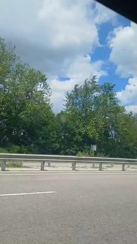 The video captures a view from a vehicle moving along a road. On the left side of the road, there are trees and green foliage. A metal guardrail separates the road from the area with trees. A street light is visible. In the distance, there is a concrete mixer truck. The sky is blue with scattered white clouds. The road appears to be asphalt.