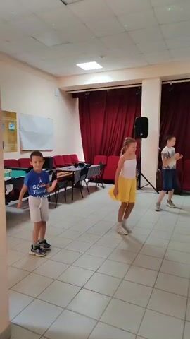 A group of children are dancing in a room with red curtains and a tiled floor. In the foreground, a boy in a blue shirt and white shorts is standing. To his right, a girl in a white top and yellow skirt is dancing. In the background, a girl in a purple shirt and white pants is standing, and another child in a striped shirt is dancing. There are speakers and chairs visible in the room. The overall impression is that of a dance class or party.