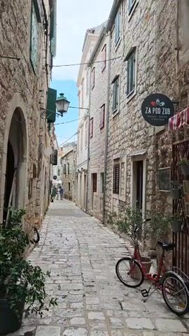 A narrow, cobblestone alleyway stretches into the distance, flanked by tall, stone buildings. The buildings have rough, weathered facades, some with green shutters and window boxes. A vintage-style streetlamp hangs from a bracket on the left building. Two bicycles, one red and one partially visible, are parked on the right side of the alley. In the distance, a few people are walking away from the camera. The sky above is a pale blue with some wispy clouds. A sign with the text "ZA POD ZUB" is visible on the right building. The overall impression is of a charming, historic European street.
