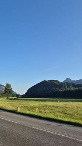 A car drives down a paved road. On the left side of the road, a grassy field stretches out towards a line of trees. Beyond the trees, rolling hills covered in lush green foliage rise up, with a few small buildings visible nestled among them. The sky above is a clear, vibrant blue, with a single wisp of a cloud. The sun appears to be low in the sky, casting long shadows and bathing the landscape in a warm, golden light, suggesting it is either sunrise or sunset. The overall impression is one of peaceful, natural beauty, with the road serving as a pathway through the serene countryside.