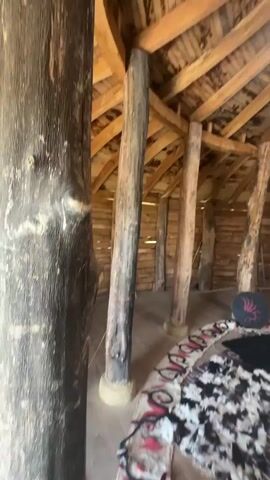 This media file presents a series of short video clips, seemingly from a personal recording, offering glimpses into an interior space with a rustic, perhaps historical, theme and then transitioning to an outdoor scene.

The interior shots focus on the architecture of a circular structure, likely a yurt or a similar dwelling. The walls are constructed of horizontal wooden logs, and the ceiling is a conical wooden framework with exposed beams. Several thick, natural wooden pillars support the roof. The floor is partially covered with animal hides, rugs with geometric patterns, and a circular dark mat with a red emblem. A circular opening in the ceiling allows natural light to illuminate the space. In one clip, a simple wooden bench is visible, along with a red and black plaid textile hanging on the wall. A dark circular object with a red emblem is also present. The overall impression of the interior is one of simple, natural materials and a cozy, perhaps primitive, living space. The scene appears to be during the daytime, with bright natural light. There are no people visible in the interior shots.

The final clip shifts to an outdoor scene. This appears to be a courtyard or an open area with a wooden deck in the foreground. Beyond the deck, a well-maintained grassy area is visible, with a stone pathway leading towards a wooden palisade fence and a gate structure. Trees and greenery are scattered around the landscape, and the sky is blue with scattered white clouds. The sun is bright, suggesting fair weather. This outdoor scene evokes a historical or reenactment setting, possibly a fortified village or a historical park. The location context provided, Neofit Rilski, Bulgaria, suggests that this might be a historical site or a place dedicated to showcasing Bulgarian history or traditions. No text is visible in any of the clips.