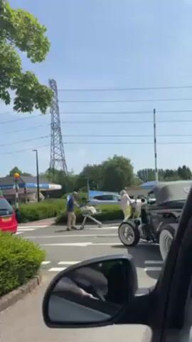 Here's a detailed description of the video:

**Scene:** The video is shot from inside a car stopped at an intersection. The time of day is daytime, with a bright, sunny sky. 

**Environment:** The location appears to be a suburban area. There's a gas station visible across the intersection, along with typical street infrastructure like traffic lights, crosswalks, and lampposts. A high-voltage power line tower stands tall in the background. Green foliage, including trees and bushes, is present.

**Objects:** 
*   **Cars:**  The primary focus is on a dark blue vintage hot rod car with wide white-walled tires. Other cars are visible in the background and to the left.
*   **Shopping Cart:** A man pushes a silver shopping cart across the crosswalk.
*   **Street Fixtures:** These include a traffic light pole, a height restriction barrier, and road signs.
*   **Reflection:** The car's side mirror is visible in the foreground, reflecting part of the interior of the car.

**People:**
*   **Man Pushing Cart:** An adult male is walking across the crosswalk, pushing a shopping cart.
*   **Hot Rod Driver:** A woman is sitting in the hot rod car, as seen from the side.
*   **Passenger:** Another person is visible in the hot rod car.
*   **Videographer:** The person recording is inside the car from which the video is shot.

**Actions:**
*   The hot rod is waiting at the intersection.
*   The man is pushing the shopping cart across the street.
*   The videographer is filming the hot rod and the intersection from within their car.

**Interactions:**
*   The hot rod waits as the man pushes the shopping cart across the street.
*   The woman in the hot rod appears to be interacting with the passenger.

**Overall Impression:** The video captures a moment of everyday life at a busy intersection, where a classic car and a man pushing a shopping cart share the road.