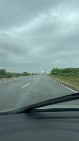 The video shows a view from inside a car looking through the windshield. The windshield wipers are visible in the foreground. The car is on a wet, paved road, indicating it is raining or has recently rained. The road is a two-lane highway with white lines marking the lanes. On either side of the road, there is vegetation and trees. In the distance, a white tanker truck is driving on the same road, heading away from the camera. There are yellow signs with red borders on the side of the road. The sky is overcast and cloudy.